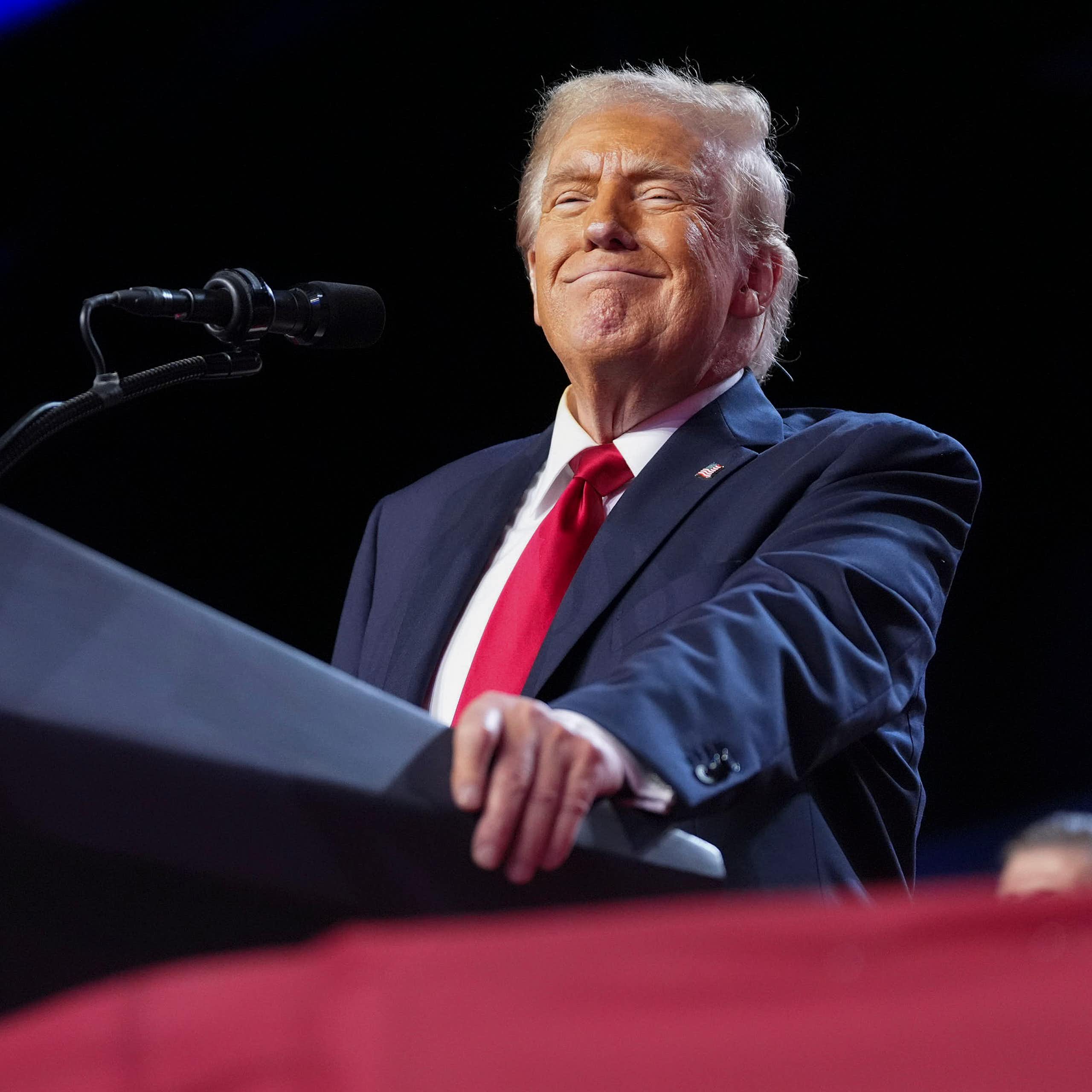 A smiling man at a lectern in a blue jacket, white shirt and red tie.