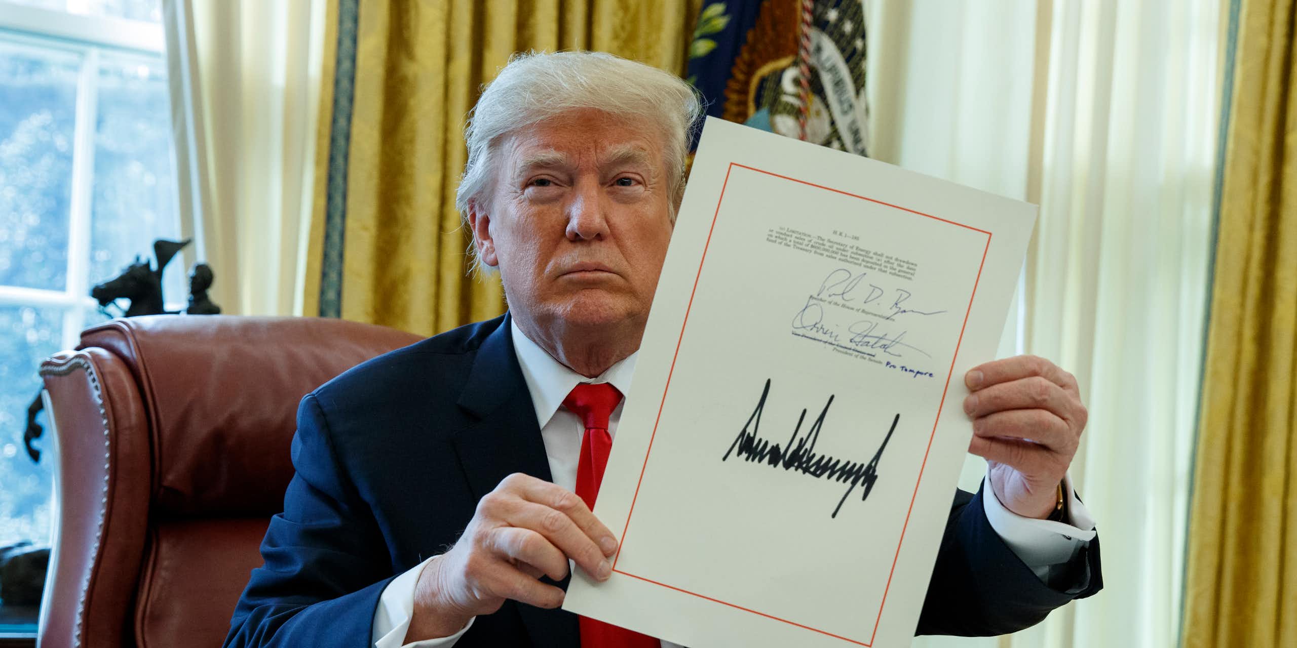 a man in a suit sitting at a desk holds up a document with his signature