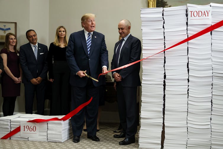 People in suits stand next to two piles of paper, while one person in a suit cuts a red ribbon