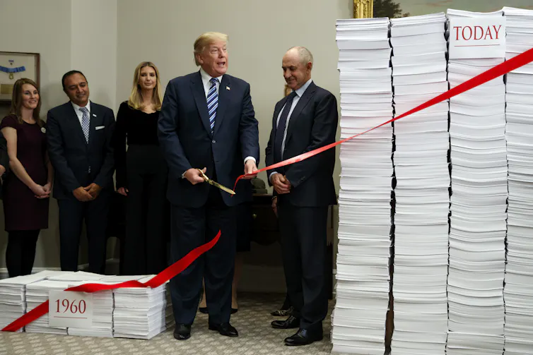 People in suits stand next to two piles of paper, while one person in a suit cuts a red ribbon