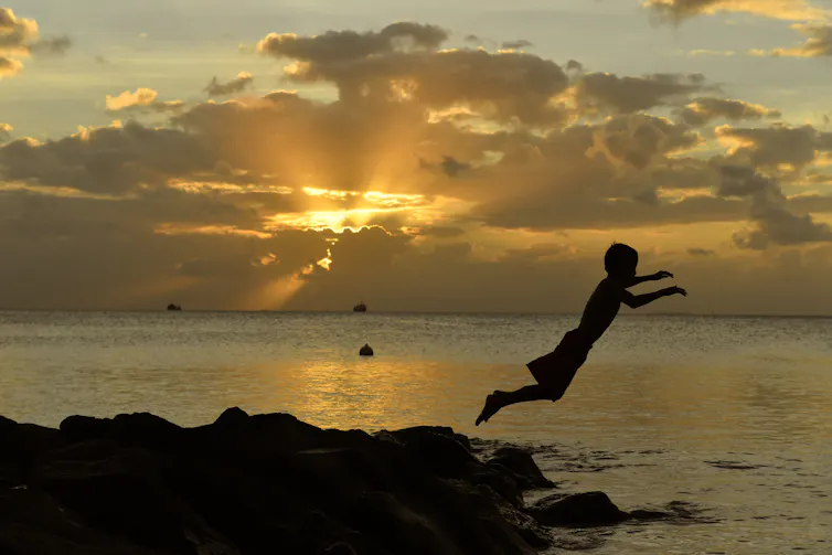 child in silhouette jumps from rock into water