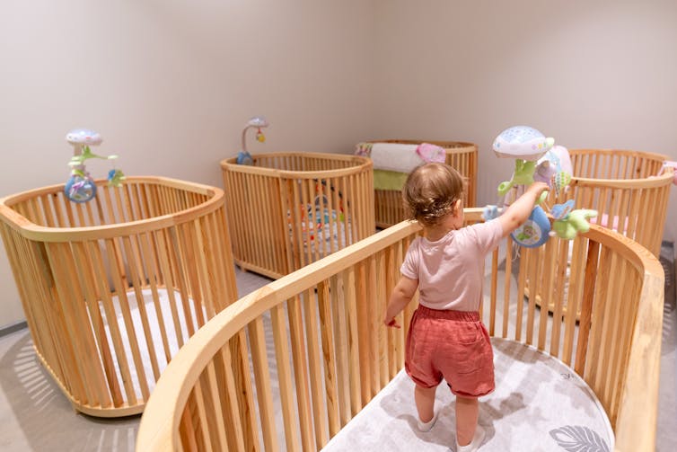 A small child standing in an oval-shaped crib touching a mobile, in a room with four other identical cribs