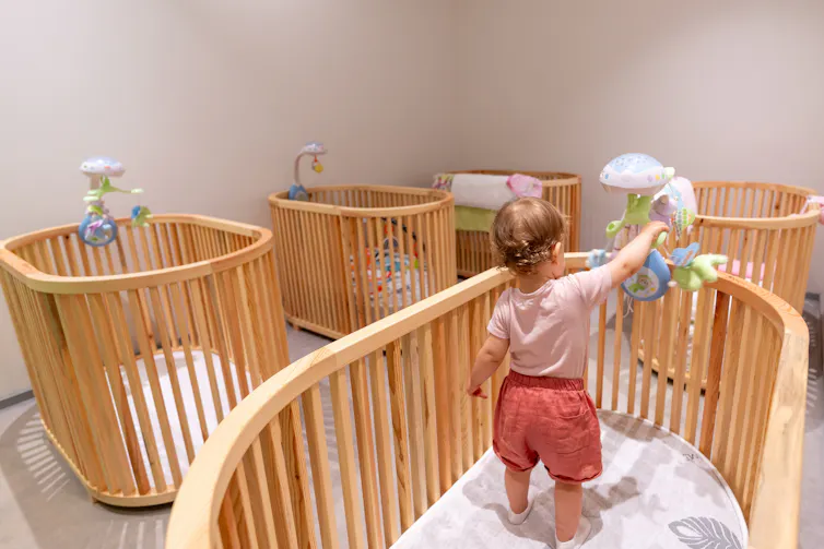 A small child standing in an oval-shaped crib touching a mobile, in a room with four other identical cribs