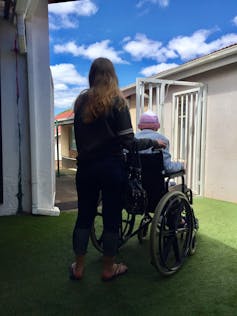 From behind, a young woman pushes an elderly person in a wheelchair, in the background blue sky and clouds.