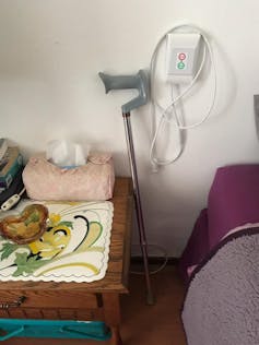 Detail of a room in an old age home, a bed and bedside table with a fluffy tissue box and a handcrafted mat, a crutch against a wall with a medical plug in the wall.