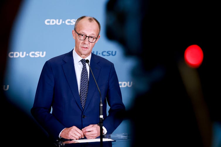 Friederich Merz stands at a podium in front of a CDU background, a video camera with a red recording light is in the foreground, obscuring part of the image