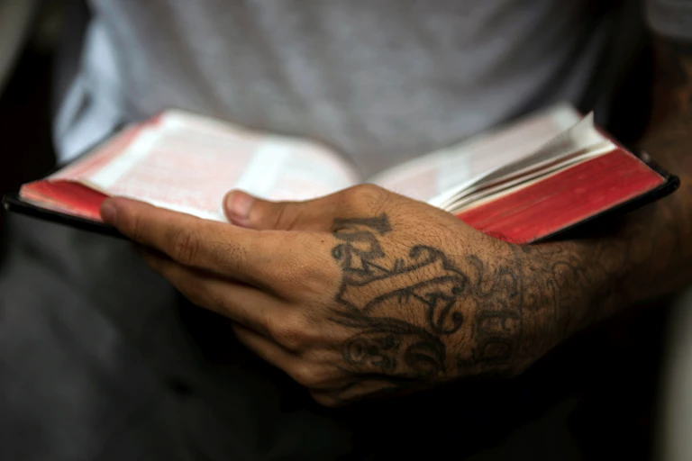 A close-up photo of a hand covered in crude tattoos holding an open bible.