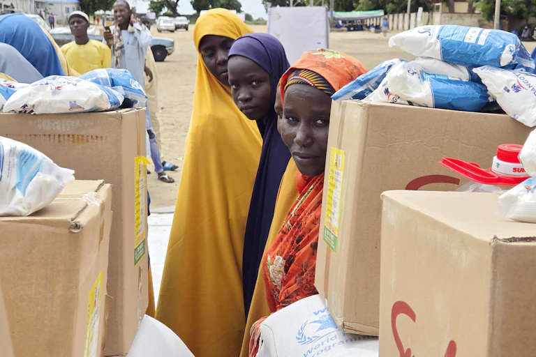 Three women in colourful garbs stand beside boxes of food supplies.