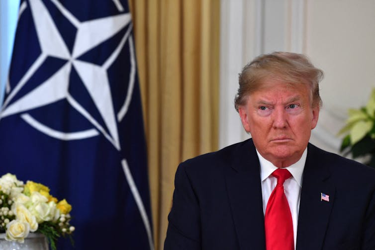 A man in a suit and red tie sits in front of a NATO blue and white flag.
