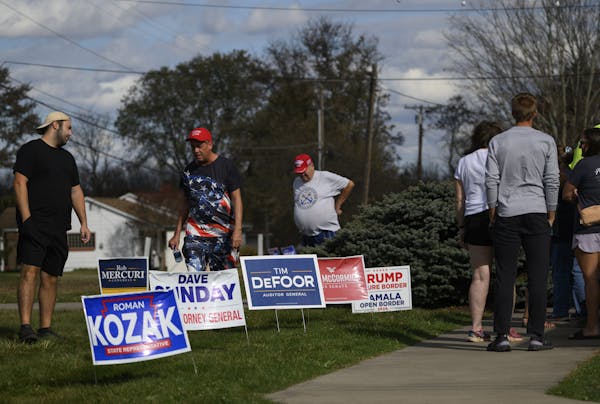 Men wearing red Trump baseball caps walk past political candidate lawn signs and people lined up to vote