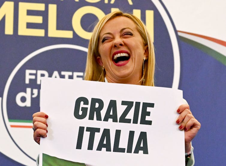 A laughing blond-haired woman holds a sign that in Italian says 'Thank you Italy.'