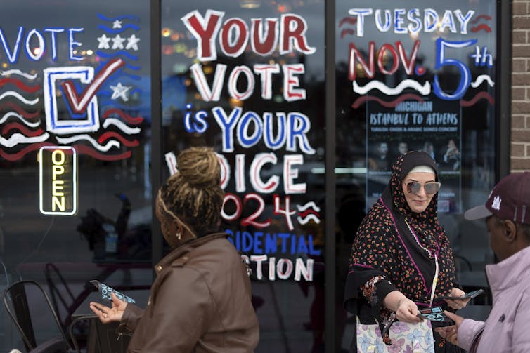 A woman wearing a head scarf and big sunglasses hands out flyers in front of a storefront painted to say