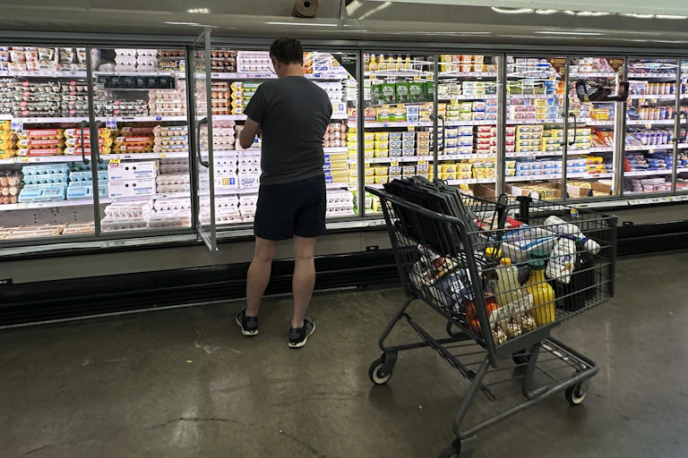 a man looks at an item in front of large grocery store fridge with many products and one door open, a grocery cart is near