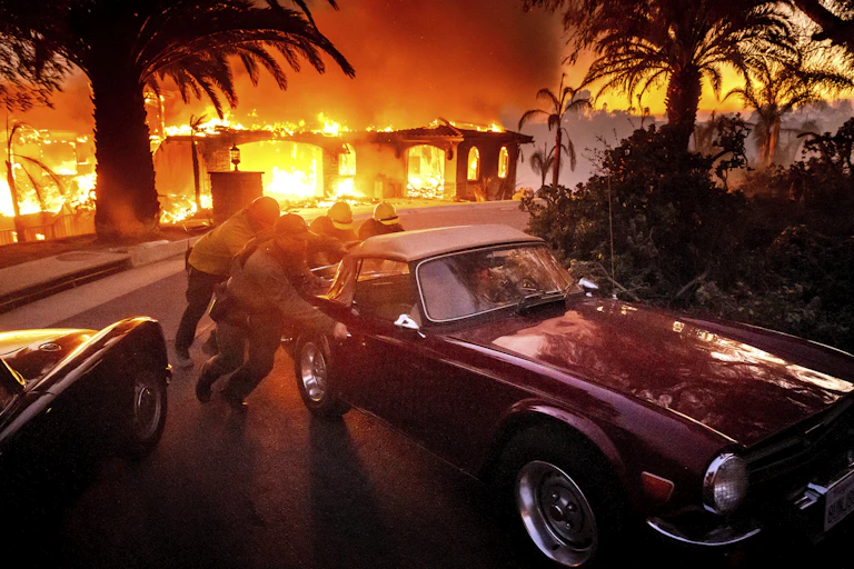 Four people push a vintage car with a canvas top away from a burning one-story house that is engulfed in flames. Palm trees are silhouetted in the yard.