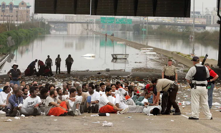 Dozens of men sit crowded together on a highway with a flooded city in the background, supervised by armed guards