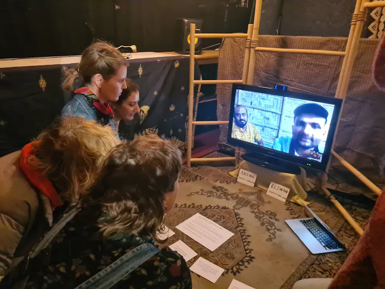 Four people crouching on a carpet in front of a screen with two men looking out at the camera on the screen. The man on the left is wearing a printed yellow shirt, the man on the right is a bit blurry and is wearing a light blue shirt and cap