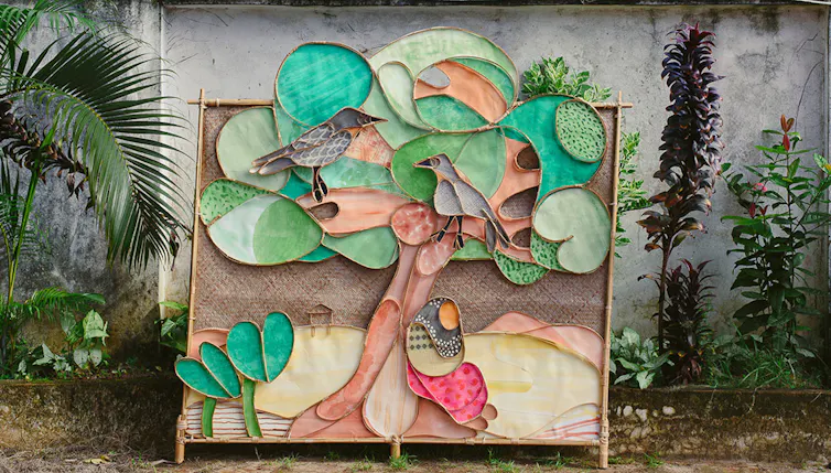 A brightly coloured story panel made from sculpted bamboo and painted elements shows two grows sitting in a banyan tree, with a women wearing a black-and-white headscarf at the tree's roots. The story panel is in front of a concrete wall with tropical plants.