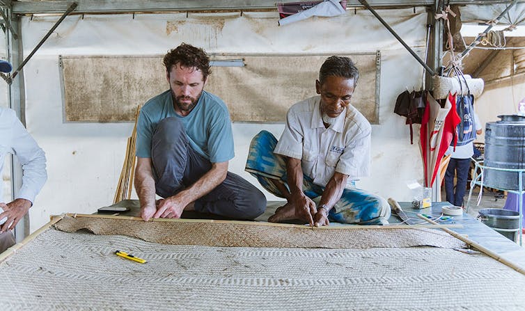 Two men sit cross-legged wearing casual shirts and long pants while handling a bamboo frame holding woven bamboo panel. They are in a large tent. There are umbrellas hanging on the right side