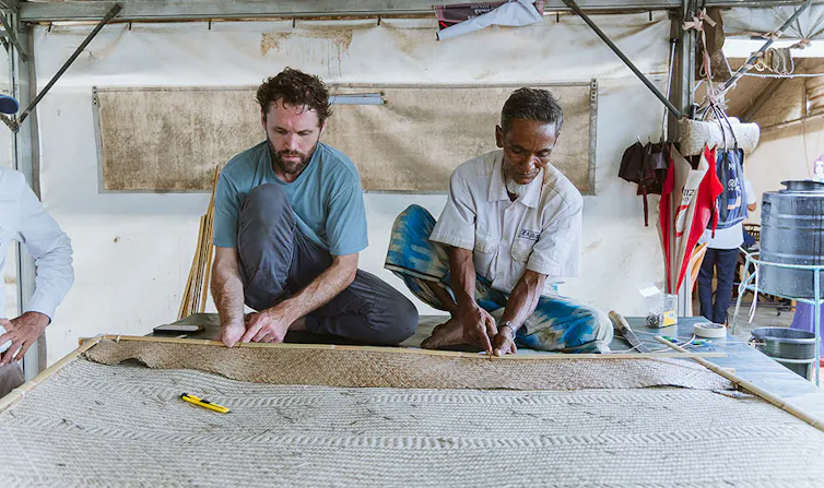 Two men sit cross-legged wearing casual shirts and long pants while handling a bamboo frame holding woven bamboo panel. They are in a large tent. There are umbrellas hanging on the right side