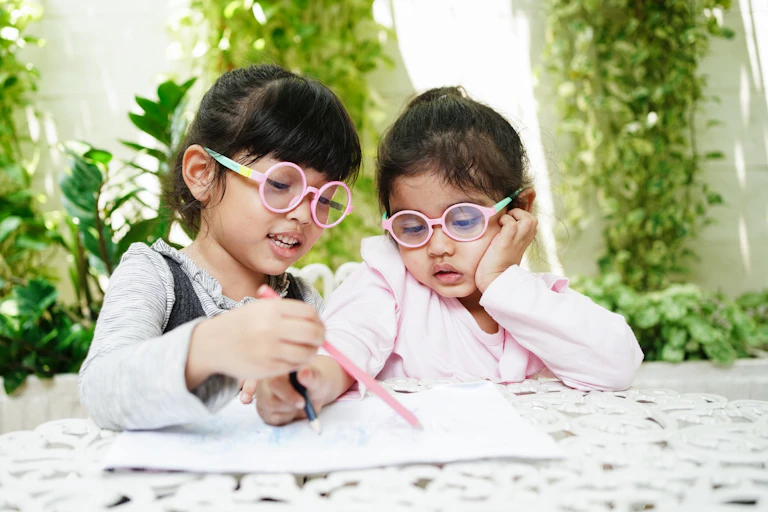 Two little girls wearing spectacles sit in a garden doing homework.