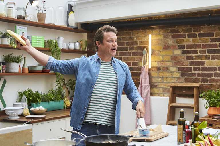 Celebity chef Jamie Oliver grates a block of cheese in a kitchen.