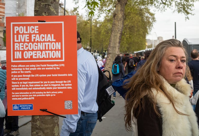 A woman walks passed an orange sign that reads: 'police live facial recognition in operation'.