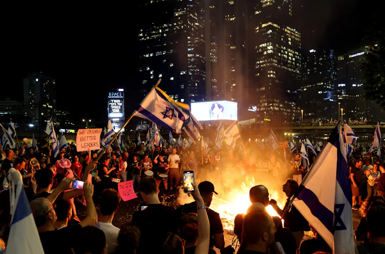 Israeli protestors gather round a fire holding signs reading 'we deserve better leaders'.