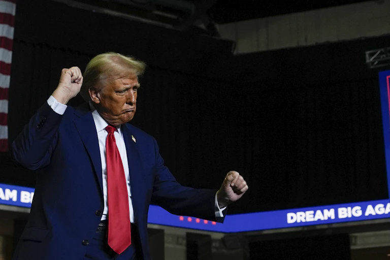 Donald trump wearing a dark blue suit and red tie dancing infront of a large sign reading dream big again