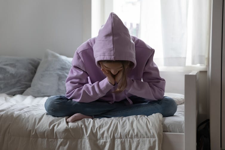 A teenage girl with her sweatshirt hooded up holds her head in her hands and stares at the floor.