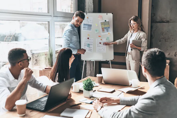 Standing woman pointing to white board while seated men listen