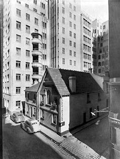 A small building with a peaked roof overshadowed by city towers.