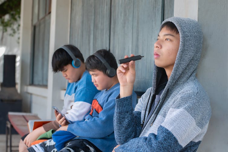 A young teenaged boy sits on a bench next to two friends wearing headphones.