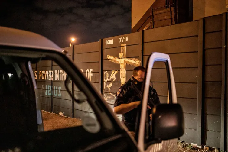A photo of a masked policeman with a gun at a car at night. Painted on a wall behind him is an image of Jesus on the cross and the words There is power in the blood of Jesus.