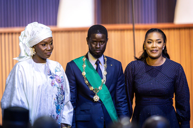 A man in a suit and tie, with a sash and a gold chain, flanked by two women, one in headscarf and robes and the other in a blue dressstanding in between two women