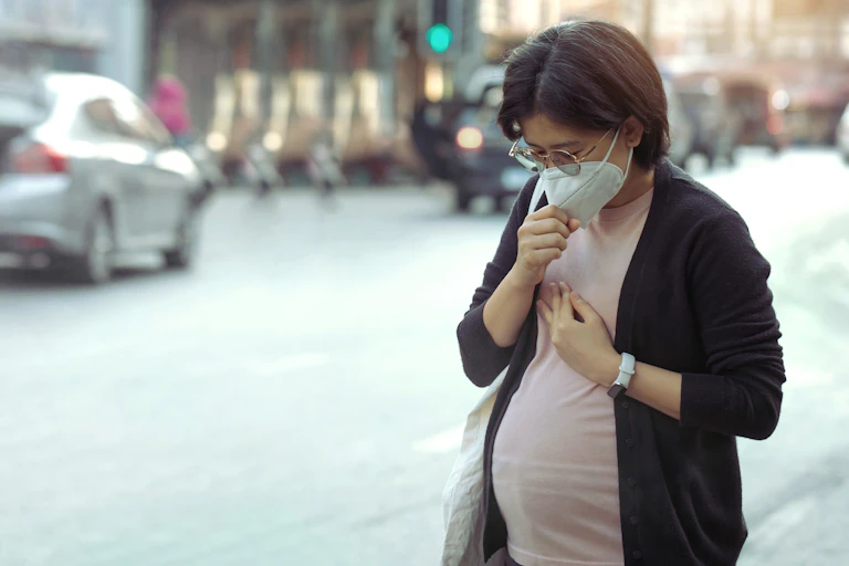 Pregnant Asian woman standing on street wearing mask, hand to her chest, traffic in background
