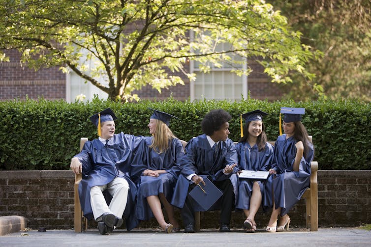Five college students in blue caps and gowns sit on a bench on campus.