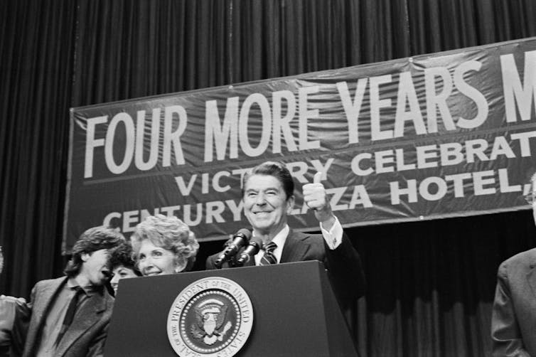 A man and woman at a podium with the presidential seal on it and a sign behind them that reads 'FOUR MORE YEARS.'
