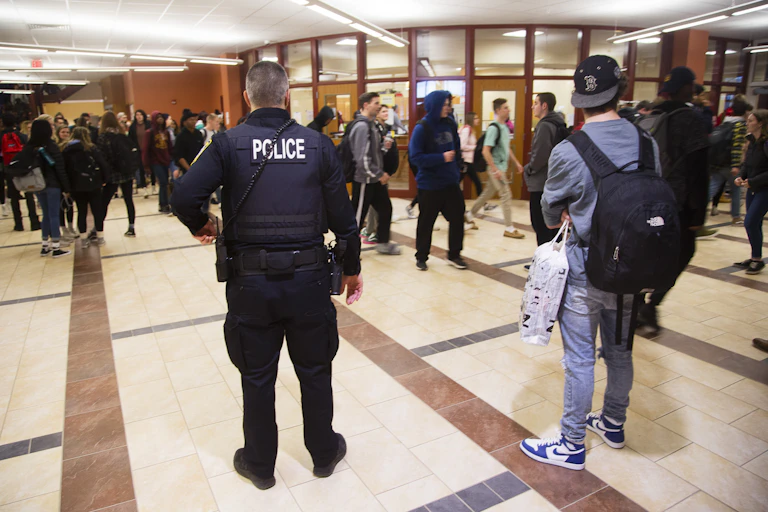 A school police officer stands in a walkway outside the main office of a high school.