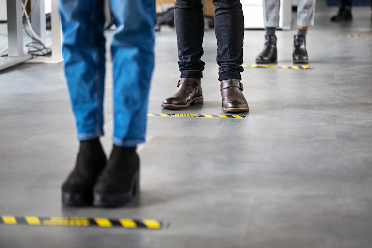 Low angle image of people standing in a queue behind social-distancing signage on the floor.