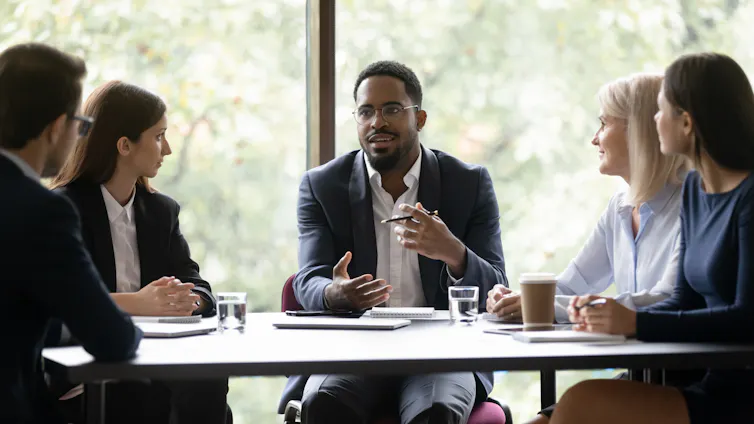 A man sits at the head of a conference table, speaking to a group of people
