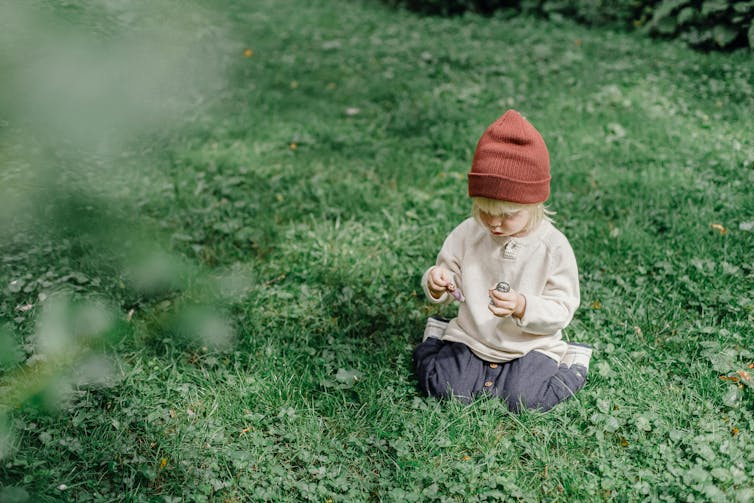 A little blonde boy in a beanie sits outside in the grass.
