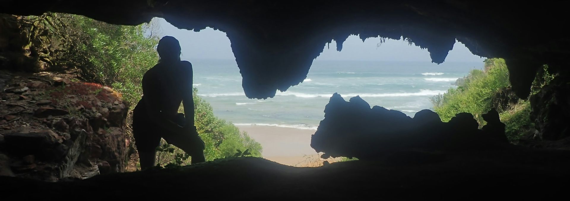 A human figure is silhouetted in the entrance to a cave, beyond which the ocean can be seen