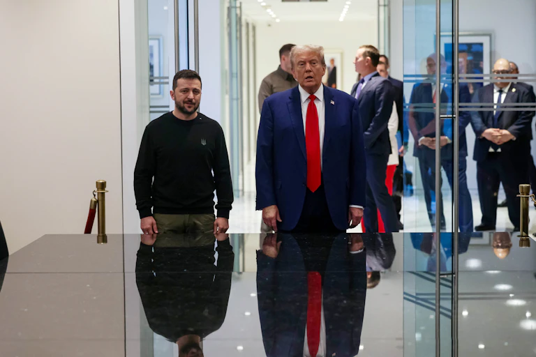 Two men, one in a blue suit the other in a sweater stand next to a table.