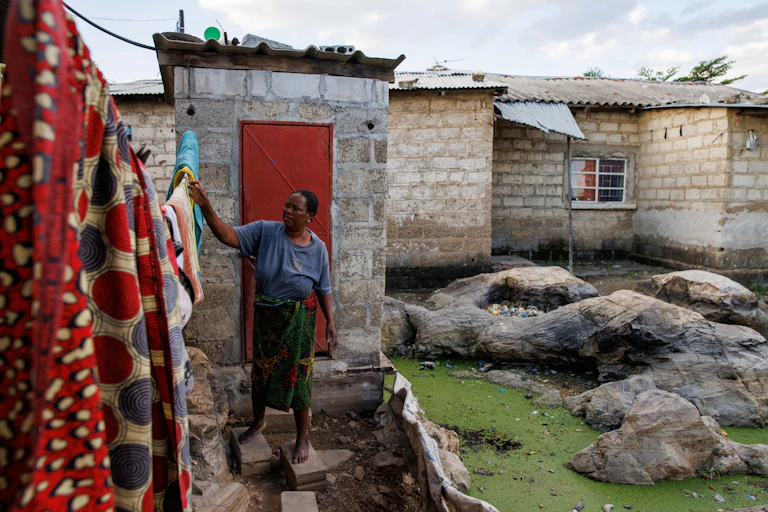 A woman feels her laundry on a washing line, next to an outdoor toilet