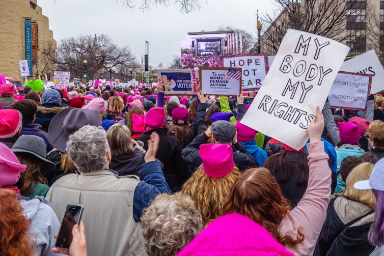 Women in pink 'pussy hats' and carrying protest signs in a march. The most visible poster in the photo reads 'my body my rights'.