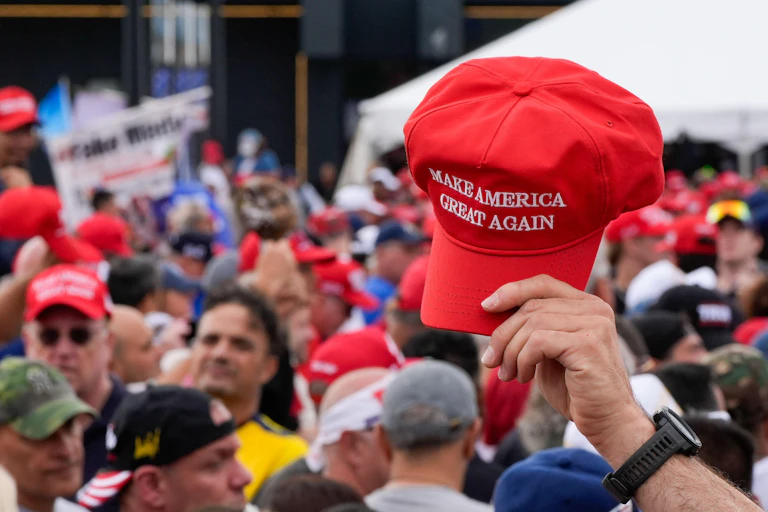 A crowd of men at a Trump rally, with one lifting a red MAGA cap above the crowd