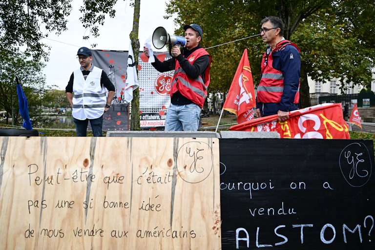 Des personnels d'Alstom en grève au moment de la vente. Ils portent des tuniques et des drapeaux CGT. Devant, des pancartes interpellent sur la vente d'Alstom à une entreprise américaine.