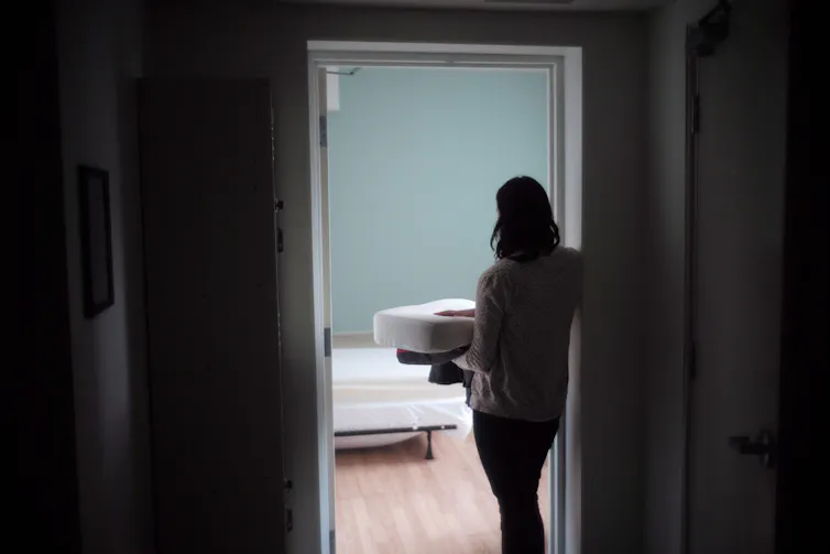 La mujer es fotografiada por detrás, de pie en la puerta de una habitación vacía con ropa de cama.
