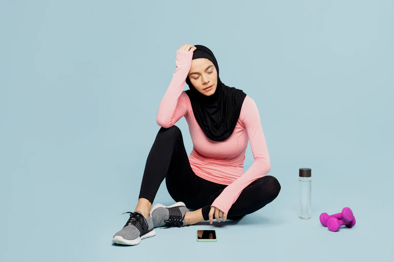 A woman wearing a hijab looks tired while sitting on the floor after a workout.
