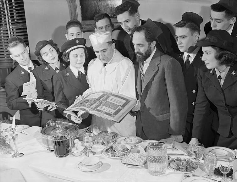 A black and white photo of young men and women in dark uniforms surrounding a pair of older men standing behind a table with a white tablecloth.